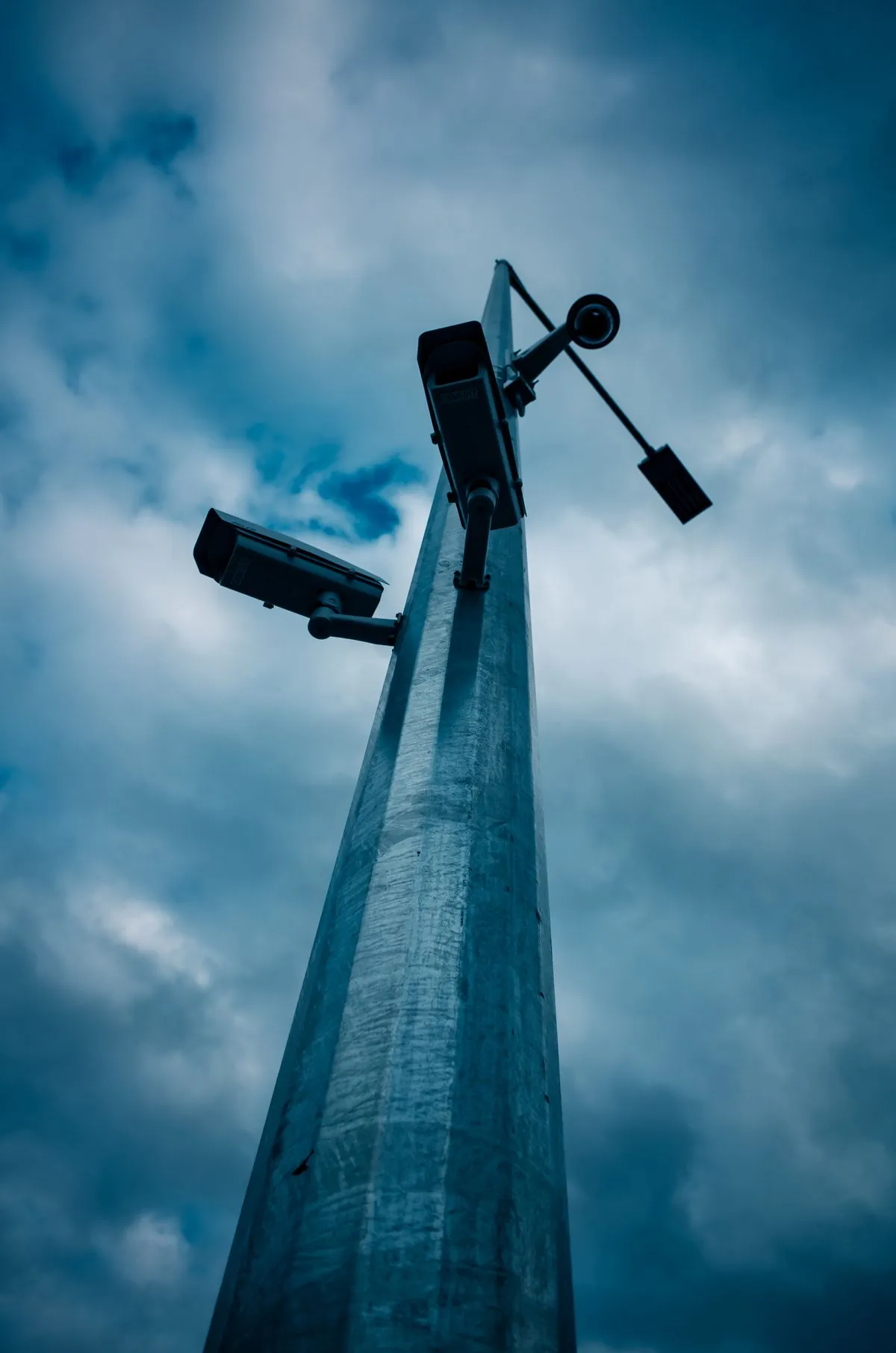 CCTV camera and floodlights mounted on a tall column against a cloudy sky